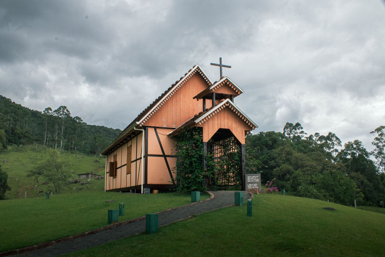 Capela de madeira em estilo enxaimel sobre um gramado, cercada de mata verde