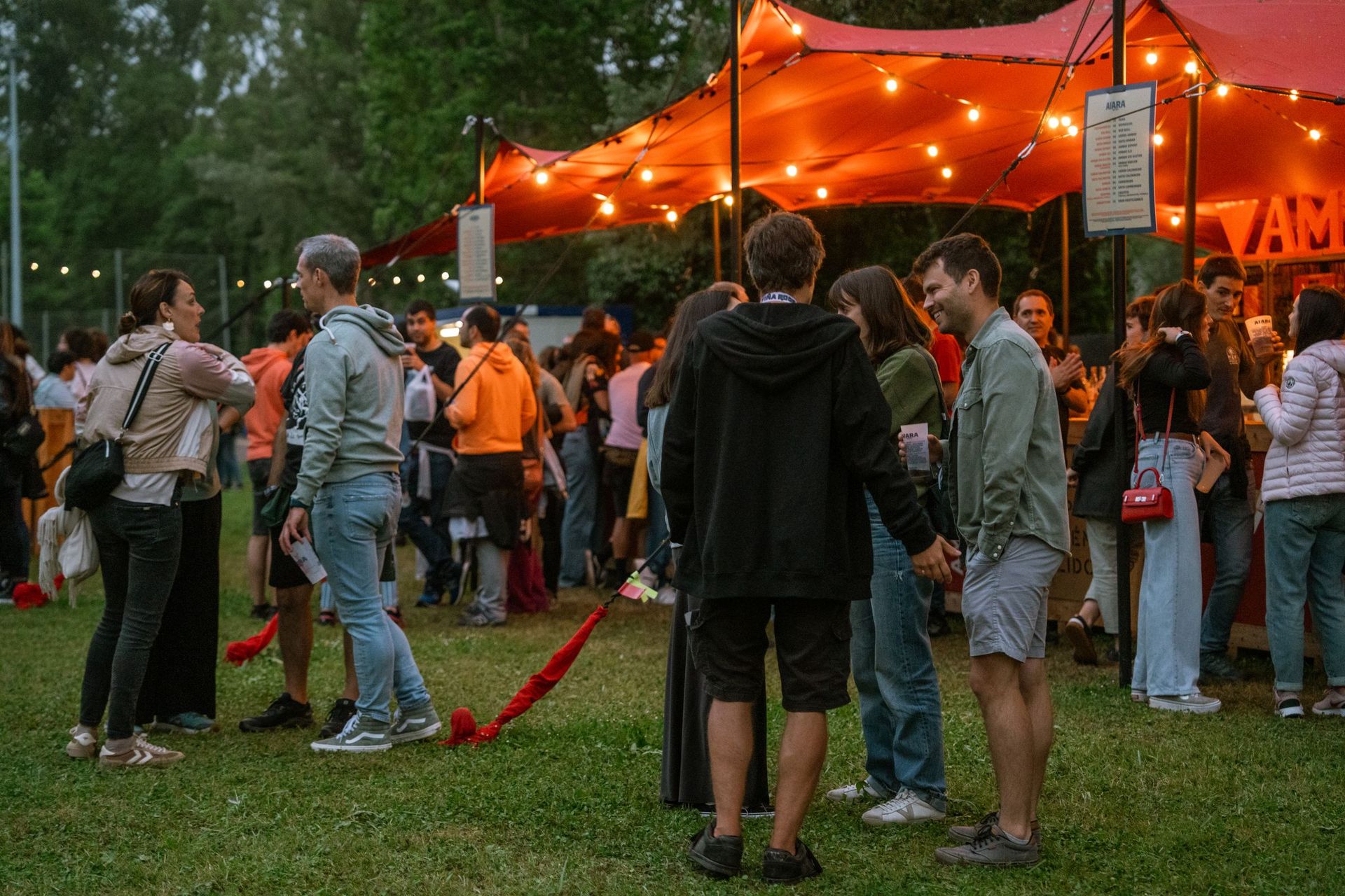 Grupo de adultos conversando no gramado de um festival ao ar livre, com tenda iluminada ao fundo