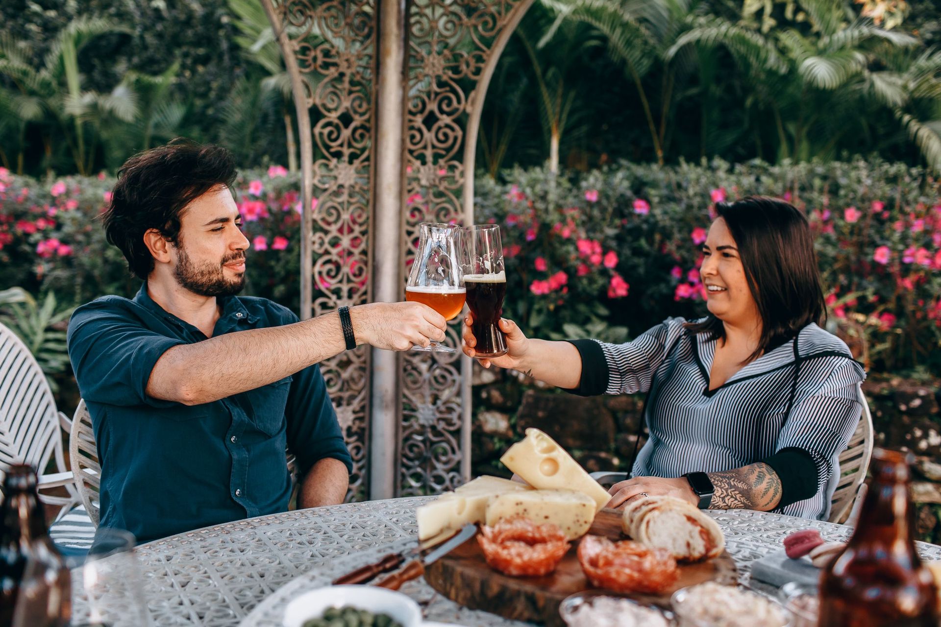 Homem e mulher brindando com copos de cerveja em uma mesa ao ar livre, cercados de plantas floridas