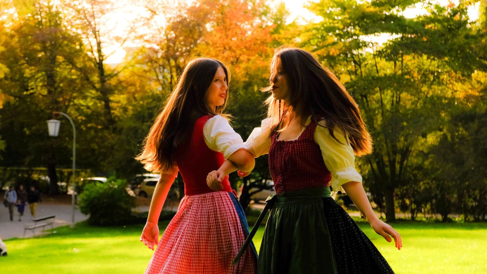 Duas mulheres com vestidos típicos (dirndl) dançando em um parque à luz do fim de tarde