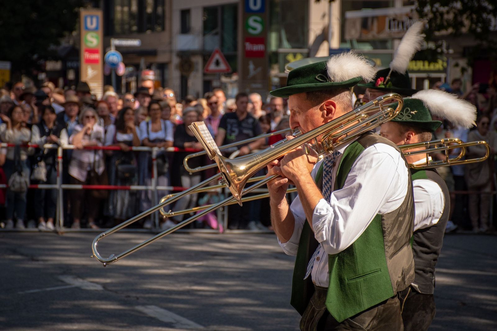 Músicos com trajes tradicionais alemães tocando trombone em um desfile de rua, com público atrás da grade