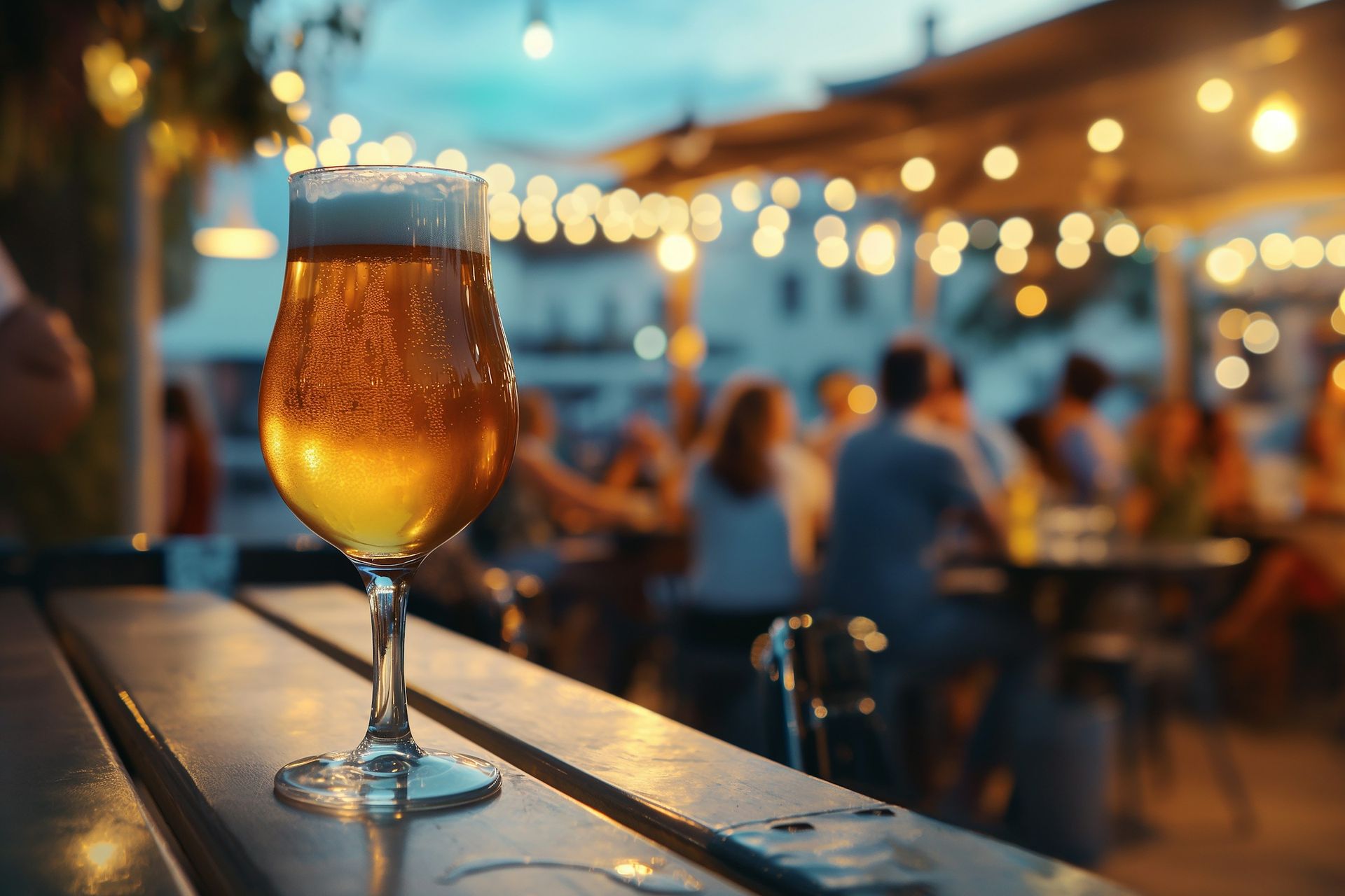 Copo de cerveja dourada sobre mesa de madeira, com varal de luzes e pessoas desfocadas ao fundo ao entardecer